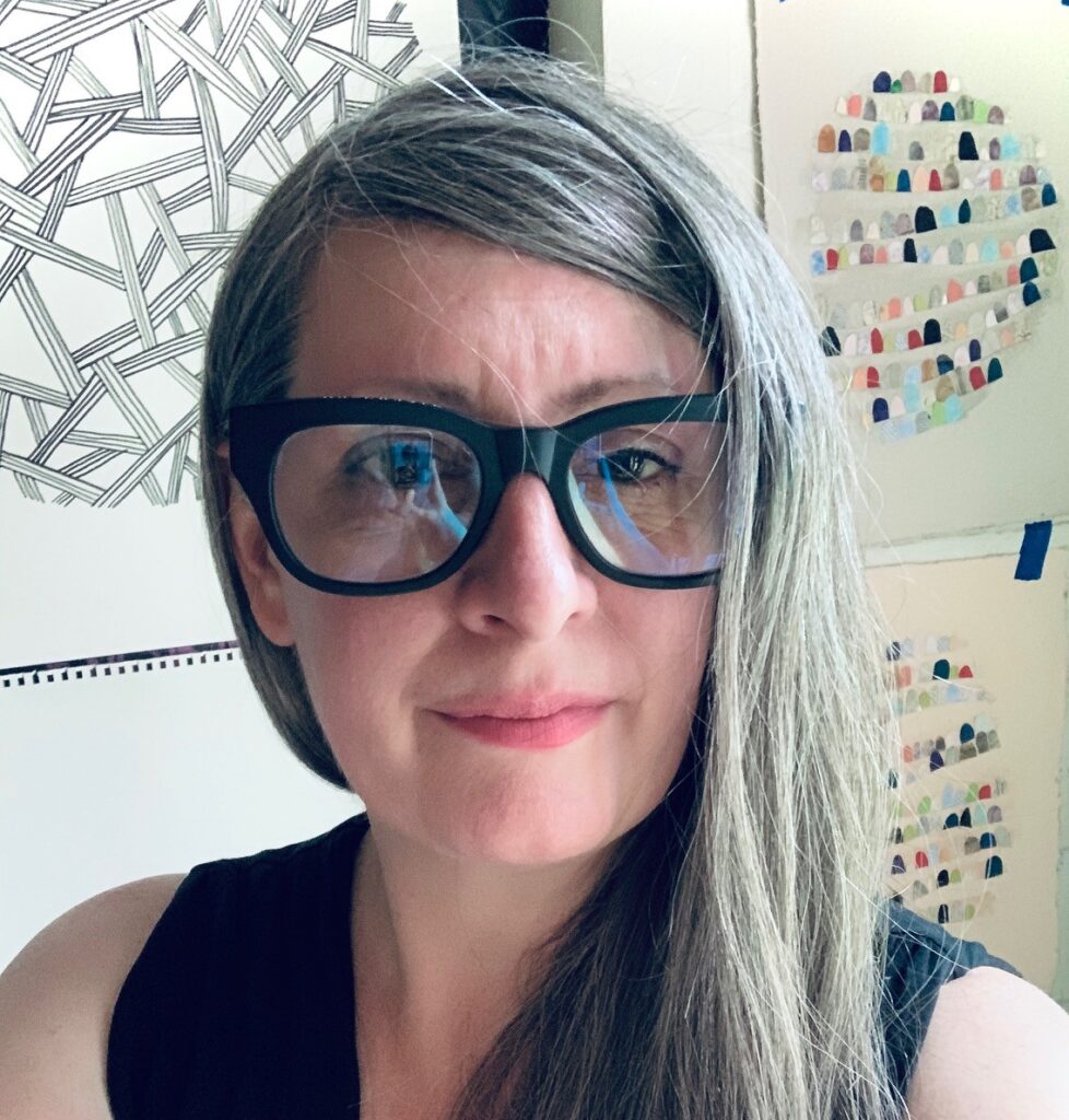 Jennifer is standing in her studio with artwork on the wall behind her. She has pale skin, brown eyes,
and long, straight brown and gray hair, parted on the side and draped over her shoulder. She is wearing
black framed glasses and a black tank.