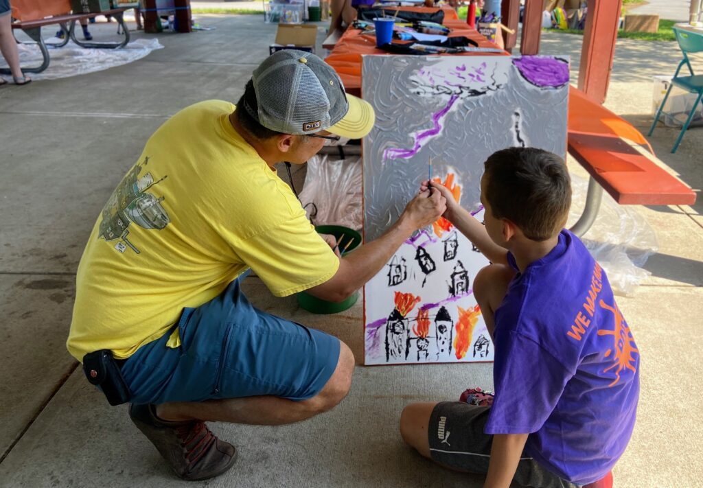A photo of two POP 2023 team participants (a father-son duo) painting on a canvas together. Both are holding paintbrushes and adding detail together.