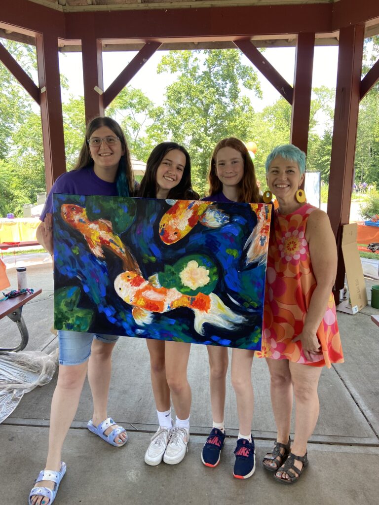 A photo of four Davis Studio team members pose with their finished painting at POP 2023. The painting is of three fish in a koi pond.