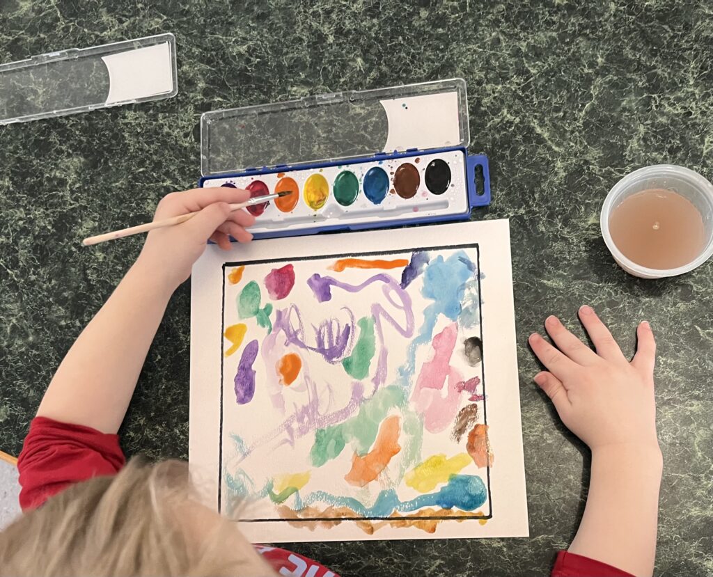A child paints with watercolors. The camera view is aerial, visible is the child's hands, the paper, and watercolors. They are painting brightly colored lines and circles.