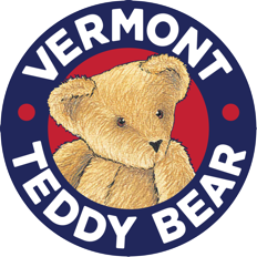 A circular logo for Vermont Teddy Bear. A navy blue circle forms the outer edge with "Vermont Teddy Bear" in white text and red dots in between the words. In the center of the circle is a brown, fuzzy teddy bear set against a red background.