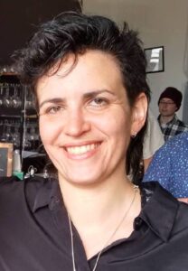 Rachel is standing in a restaurant where several people stand behind her. She is smiling and has pale skin, dark hair that is cut short, and is wearing small, silver hoop earrings. She is wearing a black collared shirt, and a necklace on a long chain.