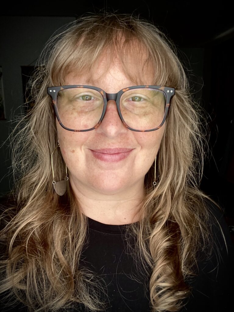 Kate is in a close up selfie. The background is black. She has blonde wavy hair with bangs and is pale skinned with freckles. She’s wearing wide tortoise shell glasses, dangling brass earrings and a solid black shirt.