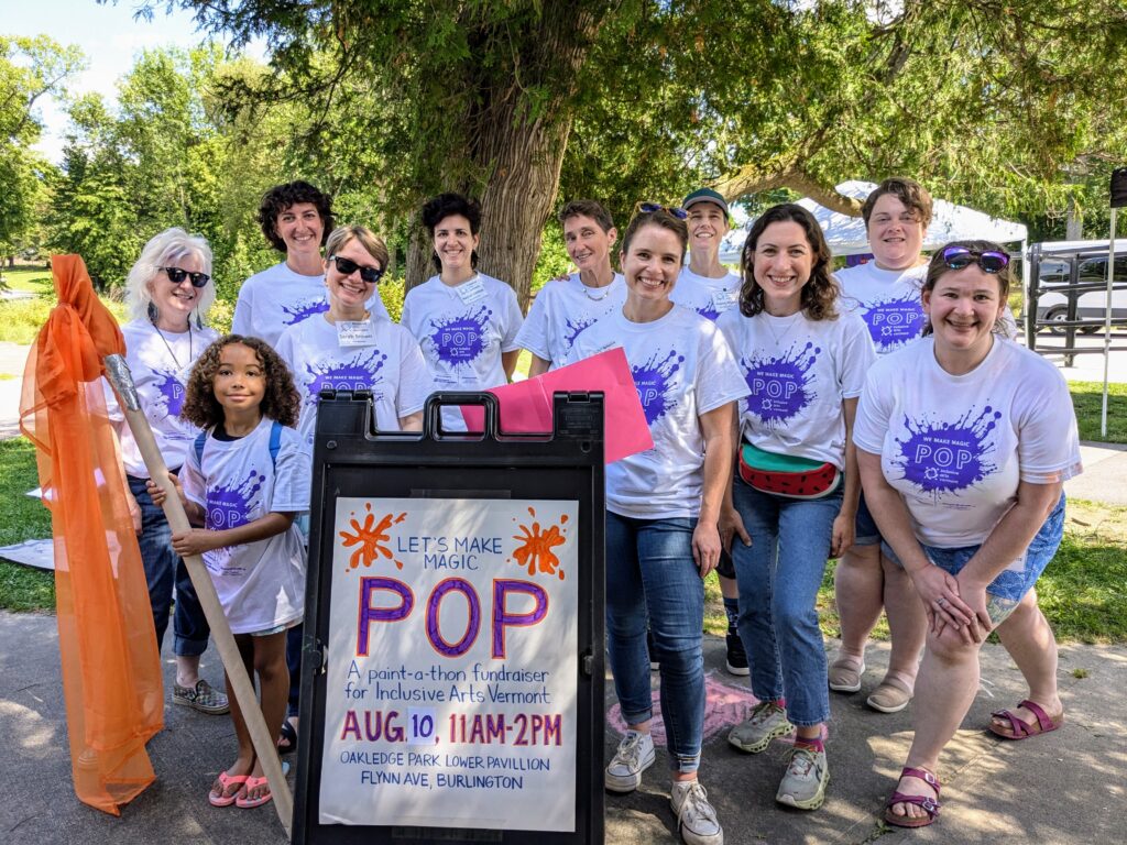 A photograph of Inclusive Arts Vermont staff and board members at the POP fundraiser outside. Everyone is smiling and wearing white t-shirts with purple paint splotches.