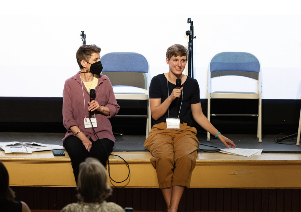 Heidi and Kirsten sit on stage together. They are each holding a microphone. Kirsten is speaking into the microphone.